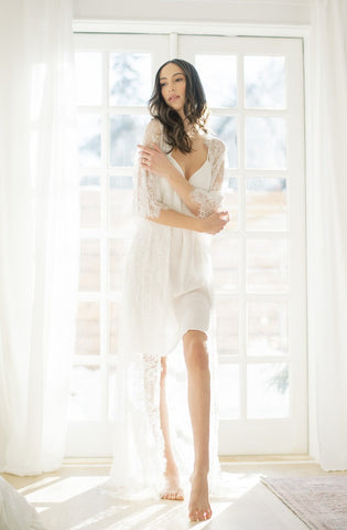 Bride standing in front of a door wearing a long white lace robe over an ivory slip dress. 