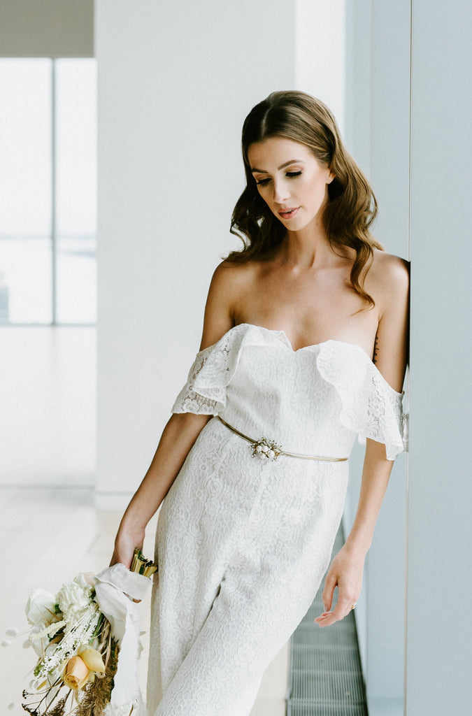 Woman leaning against wall wearing white bridal jumpsuit and holding bouquet of flowers