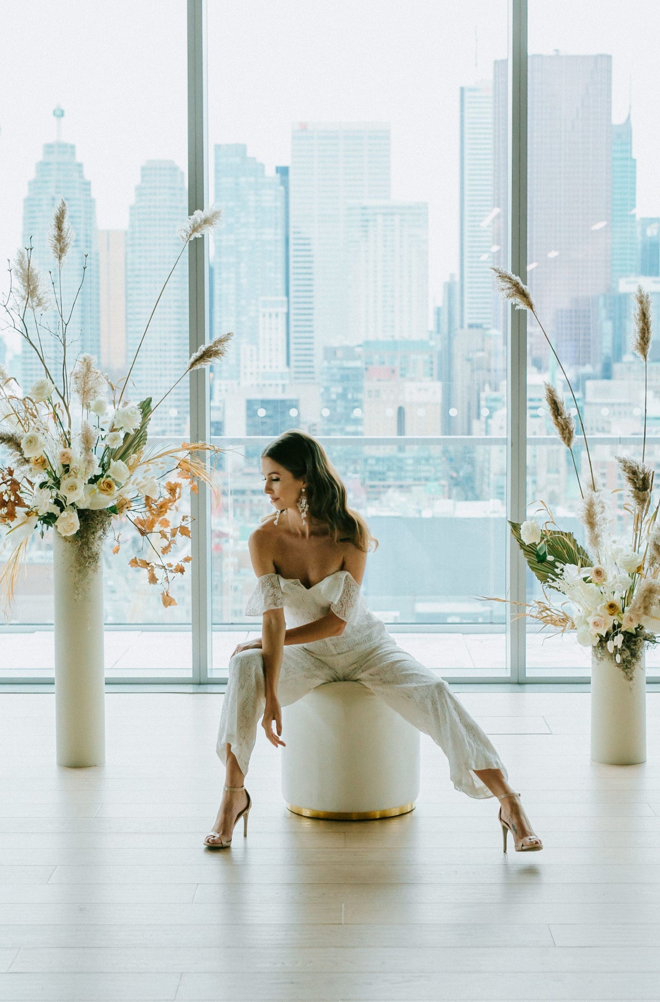 Woman sitting on stool wearing white bridal jumpsuit with cityscape behind her