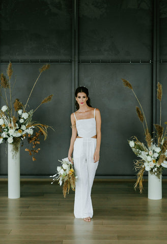 Woman walking while wearing a white bridal jumpsuit and carrying a bouquet of flowers