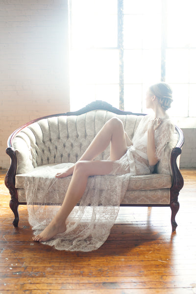 Woman looking out the window while lying on a couch wearing a long white lace robe.