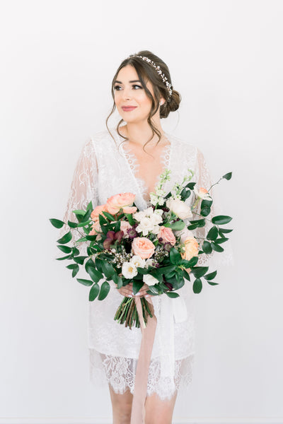 A bride holding a bouquet of flowers and wearing a white bridal robe with lace sleeves and trim. 