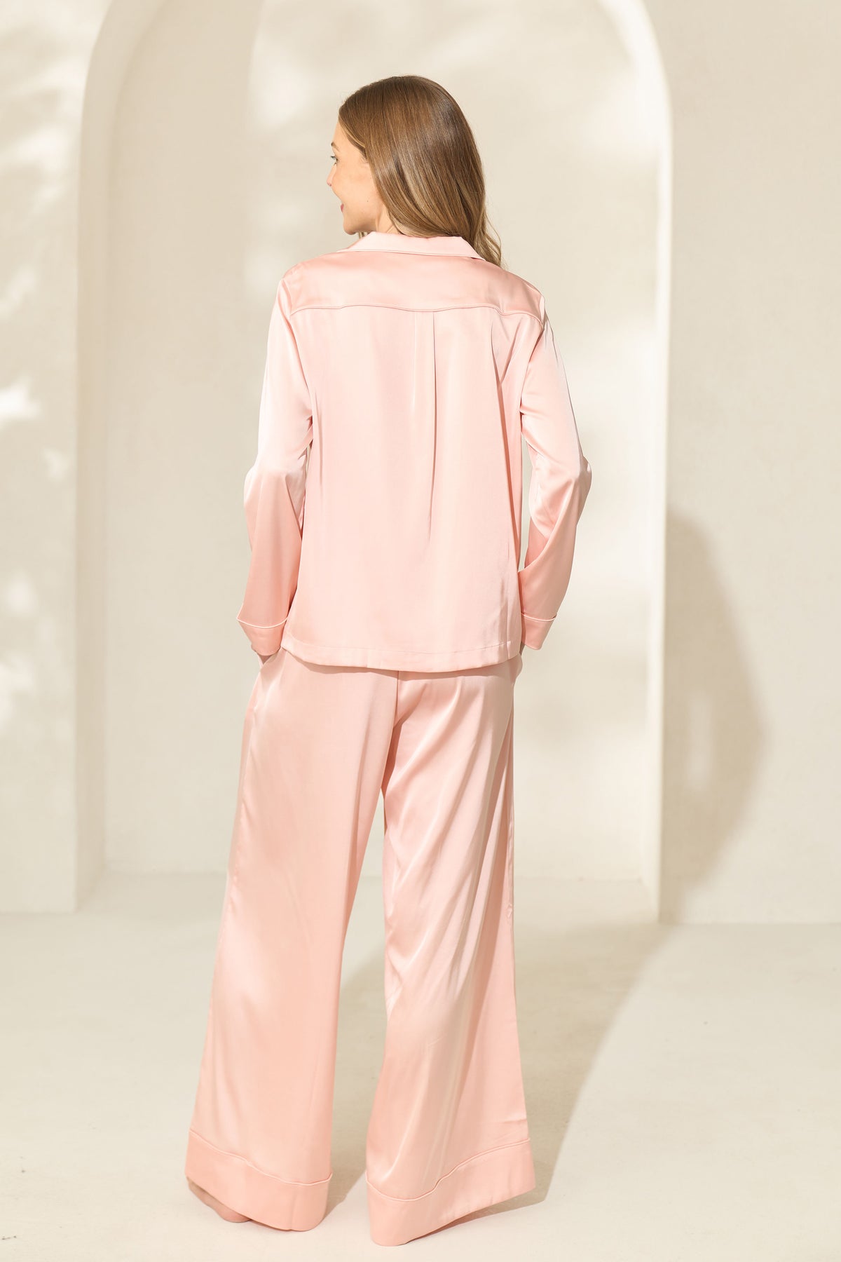 Back view of woman wearing silk-like blush pink pajama set with long sleeves. 