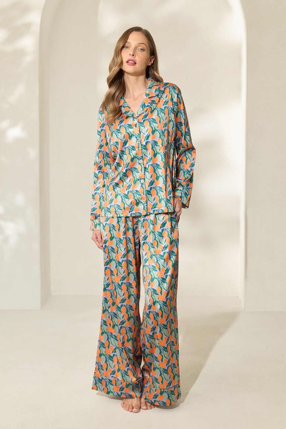 Woman wearing silk-like button-up long sleeve pajamas with green and orange citrus fruit print and elastic waistband. 