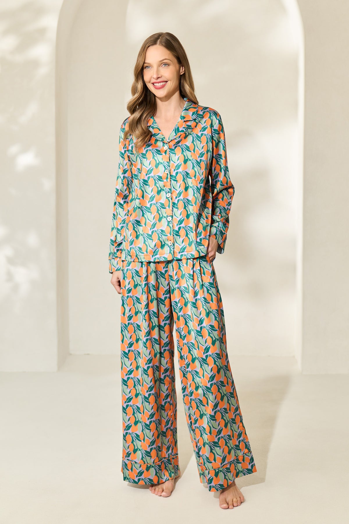 Woman wearing button-up silky long-sleeve pajama set with a green and orange citrus fruit print.