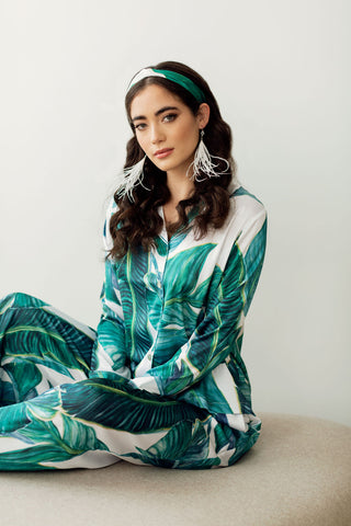 a model lounging in a chic, silky palm print pyjama set with feather earrings and matching hairband