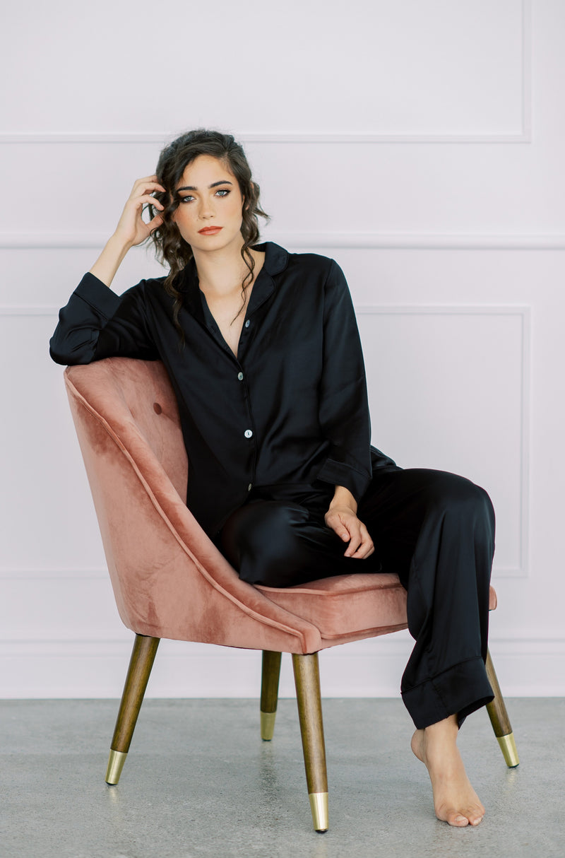 a women lounges in By Catalfo's luxurious silky black pj set, sitting in velvet chair